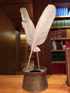 Two quill pen feathers sitting in a tin inkwell, the inkwell placed on a surface with books behind.
