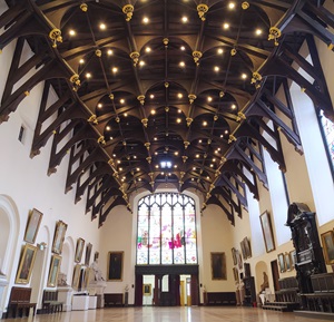 View along length of Parliament Hall, showing the dark oak timbers of the roof with their gilded points, and the stained glass window at the far end, illuminated by the sun.